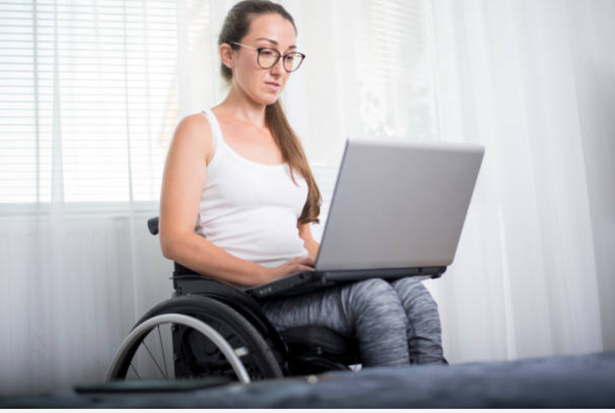 A photograph of a women sitting on a wheelchair whilst on a computer.